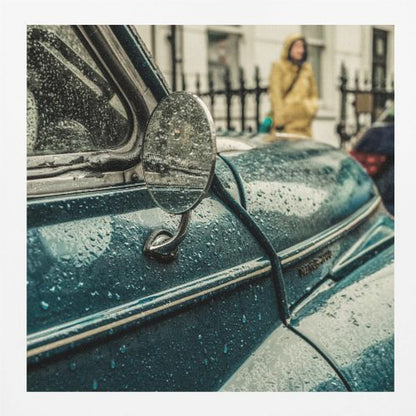 A close-up, moody photograph of a vintage teal car on a rainy day, with raindrops covering its surface. The oval side-view mirror reflects the wet street, and in the background, a person in a yellow raincoat is visible. Wall Art