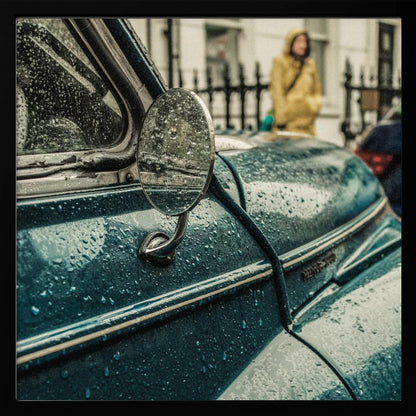 A close-up, moody photograph of a vintage teal car on a rainy day, with raindrops covering its surface. The oval side-view mirror reflects the wet street, and in the background, a person in a yellow raincoat is visible. Wall Art