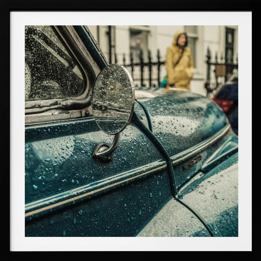 A close-up, moody photograph of a vintage teal car on a rainy day, with raindrops covering its surface. The oval side-view mirror reflects the wet street, and in the background, a person in a yellow raincoat is visible. Wall Art