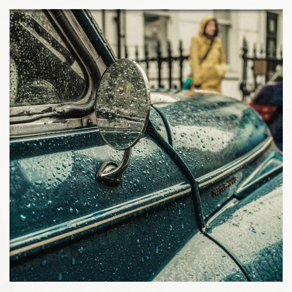 A close-up, moody photograph of a vintage teal car on a rainy day, with raindrops covering its surface. The oval side-view mirror reflects the wet street, and in the background, a person in a yellow raincoat is visible. Wall Art