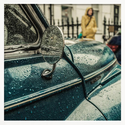 A close-up, moody photograph of a vintage teal car on a rainy day, with raindrops covering its surface. The oval side-view mirror reflects the wet street, and in the background, a person in a yellow raincoat is visible. Wall Art