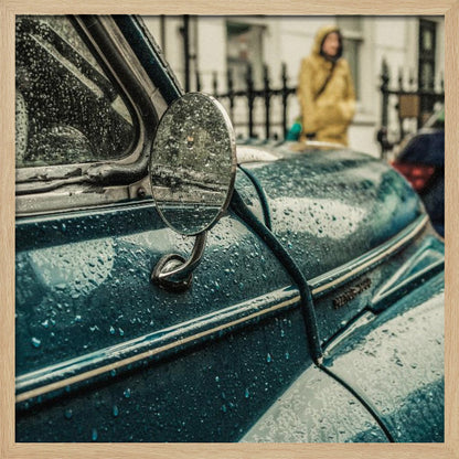A close-up, moody photograph of a vintage teal car on a rainy day, with raindrops covering its surface. The oval side-view mirror reflects the wet street, and in the background, a person in a yellow raincoat is visible. Wall Art
