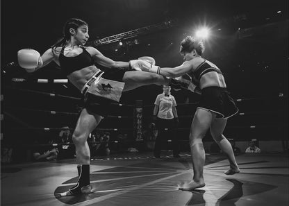 A dramatic black and white action photograph of two female kickboxers in a ring. One fighter, airborne, executes a high kick that connects with the other fighter's gloves, who is bracing for the impact. A referee stands in the background under bright arena lights. Poster