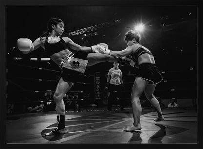 A dramatic black and white action photograph of two female kickboxers in a ring. One fighter, airborne, executes a high kick that connects with the other fighter's gloves, who is bracing for the impact. A referee stands in the background under bright arena lights. Poster