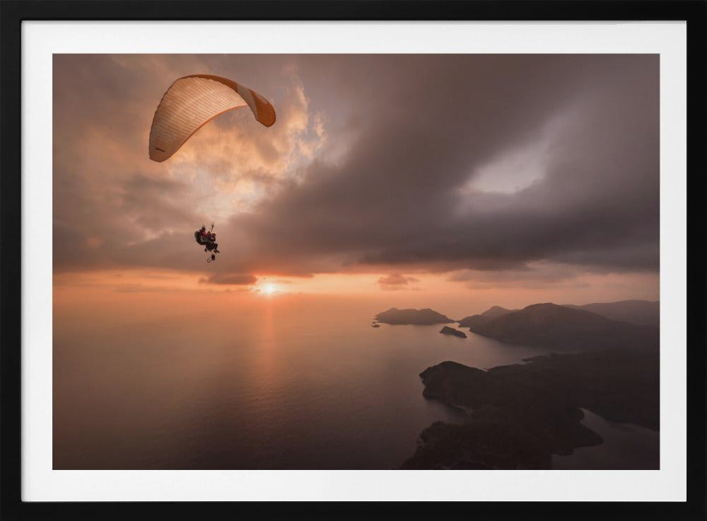 A scenic aerial view of a person paragliding over the ocean during a dramatic sunset. The orange and white parachute is in the upper left corner against a cloudy sky. The sun on the horizon casts a warm glow on the water, with silhouetted islands in the distance. Poster
