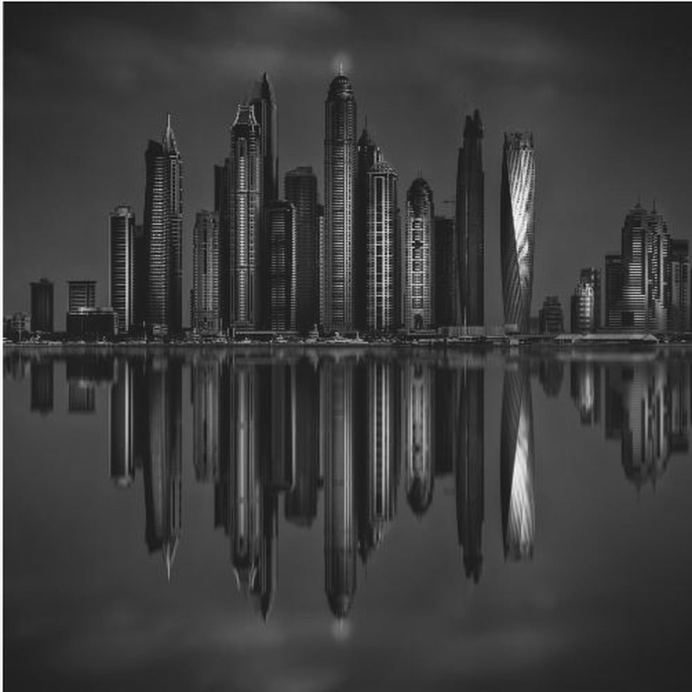 A dramatic black and white photograph of a modern city skyline at night, its towering skyscrapers perfectly reflected in the calm water below, all enclosed within a light brown wooden frame. Print