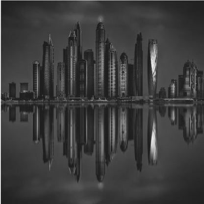 A dramatic black and white photograph of a modern city skyline at night, its towering skyscrapers perfectly reflected in the calm water below, all enclosed within a light brown wooden frame. Print