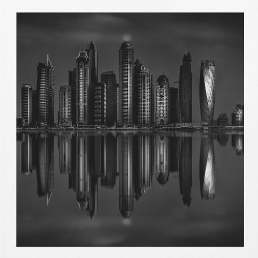 A dramatic black and white photograph of a modern city skyline at night, its towering skyscrapers perfectly reflected in the calm water below, all enclosed within a light brown wooden frame. Print