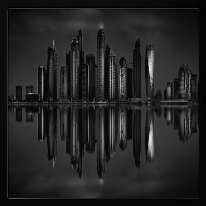 A dramatic black and white photograph of a modern city skyline at night, its towering skyscrapers perfectly reflected in the calm water below, all enclosed within a light brown wooden frame. Print