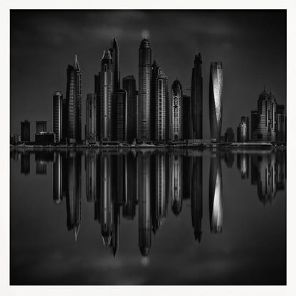 A dramatic black and white photograph of a modern city skyline at night, its towering skyscrapers perfectly reflected in the calm water below, all enclosed within a light brown wooden frame. Print