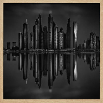 A dramatic black and white photograph of a modern city skyline at night, its towering skyscrapers perfectly reflected in the calm water below, all enclosed within a light brown wooden frame. Print
