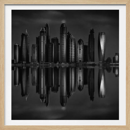 A dramatic black and white photograph of a modern city skyline at night, its towering skyscrapers perfectly reflected in the calm water below, all enclosed within a light brown wooden frame. Print