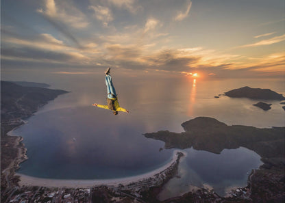 A breathtaking aerial view of a skydiver in a head-down position, freefalling over a vast coastal bay at sunset. The setting sun casts a golden glow over the ocean and clouds, highlighting the islands and coastline below. Decor