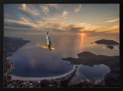 A breathtaking aerial view of a skydiver in a head-down position, freefalling over a vast coastal bay at sunset. The setting sun casts a golden glow over the ocean and clouds, highlighting the islands and coastline below. Decor