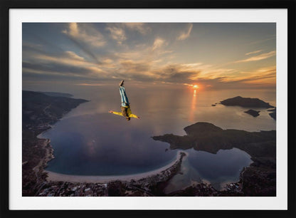 A breathtaking aerial view of a skydiver in a head-down position, freefalling over a vast coastal bay at sunset. The setting sun casts a golden glow over the ocean and clouds, highlighting the islands and coastline below. Decor