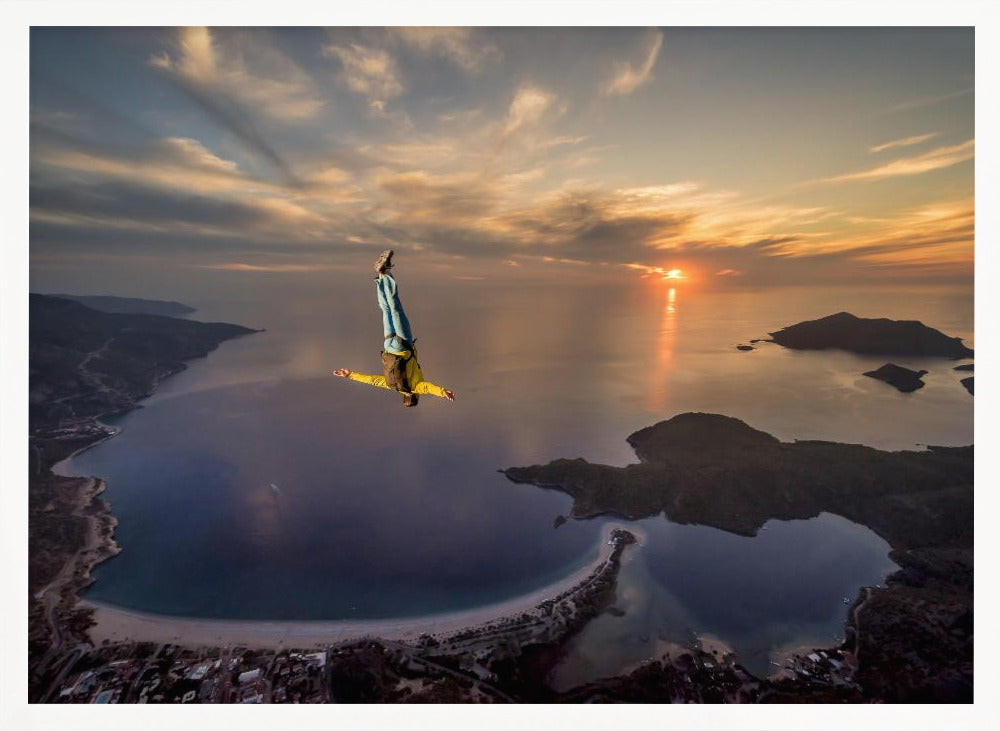 A breathtaking aerial view of a skydiver in a head-down position, freefalling over a vast coastal bay at sunset. The setting sun casts a golden glow over the ocean and clouds, highlighting the islands and coastline below. Decor