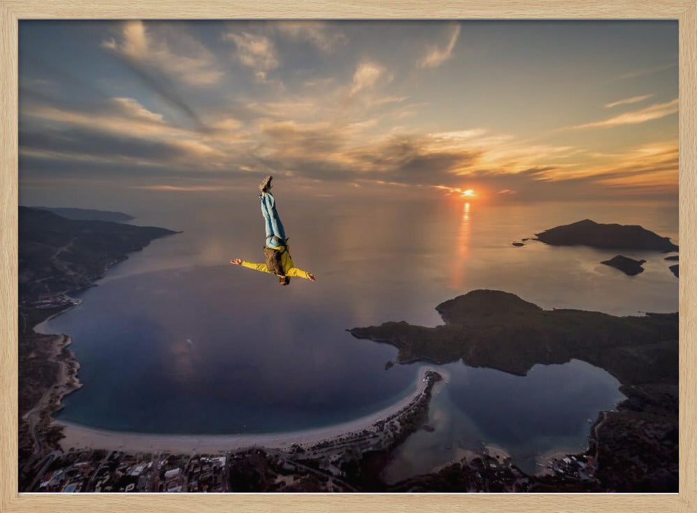 A breathtaking aerial view of a skydiver in a head-down position, freefalling over a vast coastal bay at sunset. The setting sun casts a golden glow over the ocean and clouds, highlighting the islands and coastline below. Decor