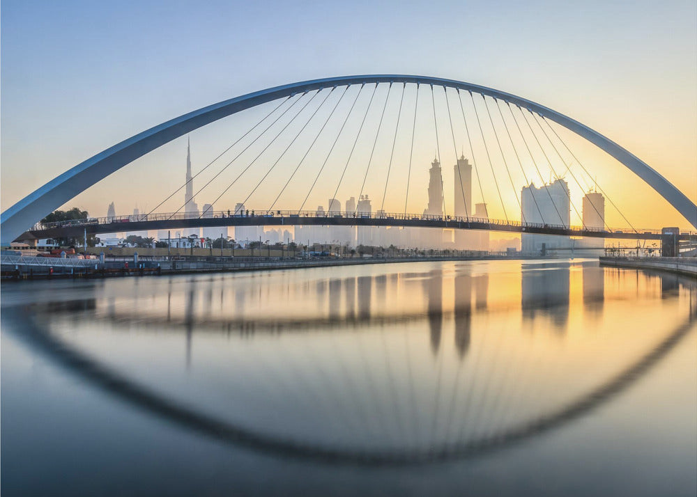 A framed photograph of the Dubai Water Canal Bridge at sunrise, with its large arch spanning across the water. The Dubai skyline, including the Burj Khalifa, is visible in the background, silhouetted against a golden and blue sky. The bridge and buildings create a perfect reflection on the calm surface of the water. Wall Art