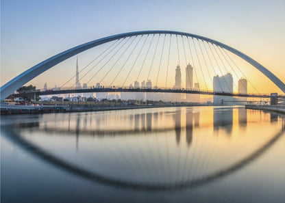 A framed photograph of the Dubai Water Canal Bridge at sunrise, with its large arch spanning across the water. The Dubai skyline, including the Burj Khalifa, is visible in the background, silhouetted against a golden and blue sky. The bridge and buildings create a perfect reflection on the calm surface of the water. Wall Art