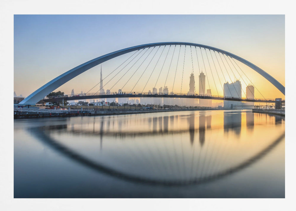 A framed photograph of the Dubai Water Canal Bridge at sunrise, with its large arch spanning across the water. The Dubai skyline, including the Burj Khalifa, is visible in the background, silhouetted against a golden and blue sky. The bridge and buildings create a perfect reflection on the calm surface of the water. Wall Art