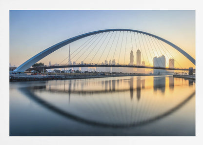 A framed photograph of the Dubai Water Canal Bridge at sunrise, with its large arch spanning across the water. The Dubai skyline, including the Burj Khalifa, is visible in the background, silhouetted against a golden and blue sky. The bridge and buildings create a perfect reflection on the calm surface of the water. Wall Art