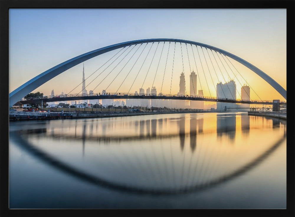 A framed photograph of the Dubai Water Canal Bridge at sunrise, with its large arch spanning across the water. The Dubai skyline, including the Burj Khalifa, is visible in the background, silhouetted against a golden and blue sky. The bridge and buildings create a perfect reflection on the calm surface of the water. Wall Art