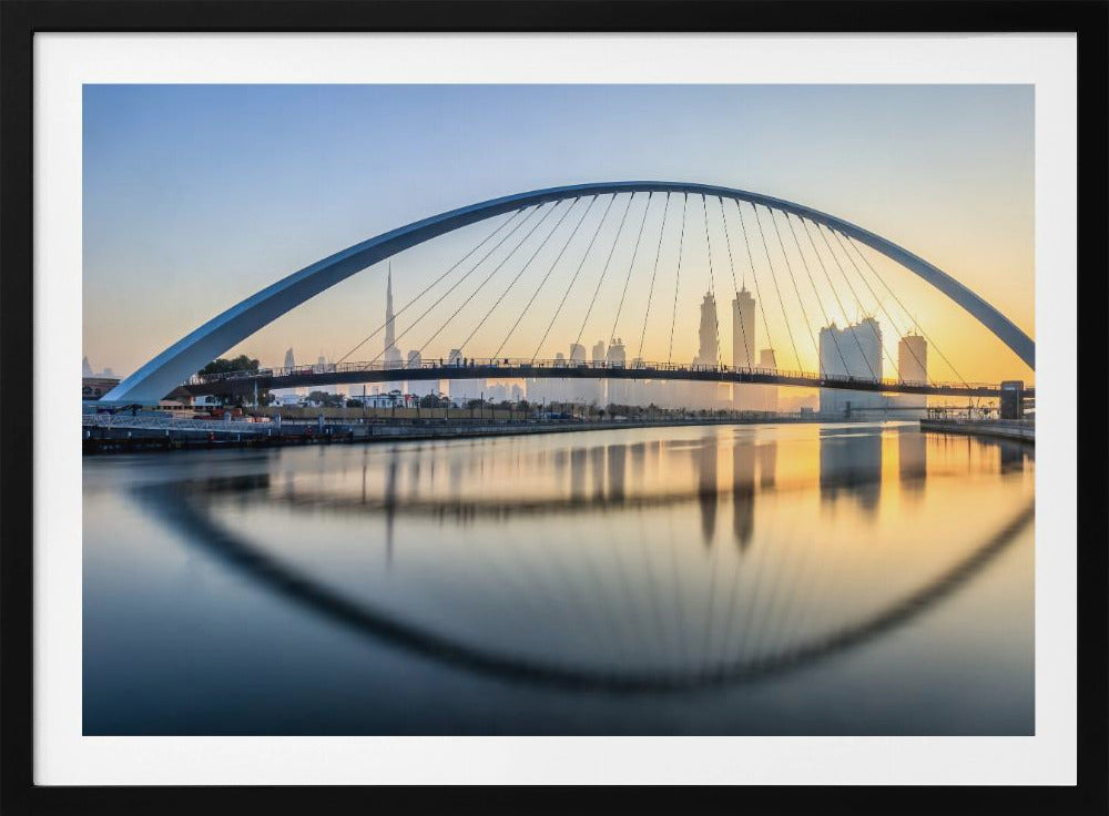 A framed photograph of the Dubai Water Canal Bridge at sunrise, with its large arch spanning across the water. The Dubai skyline, including the Burj Khalifa, is visible in the background, silhouetted against a golden and blue sky. The bridge and buildings create a perfect reflection on the calm surface of the water. Wall Art