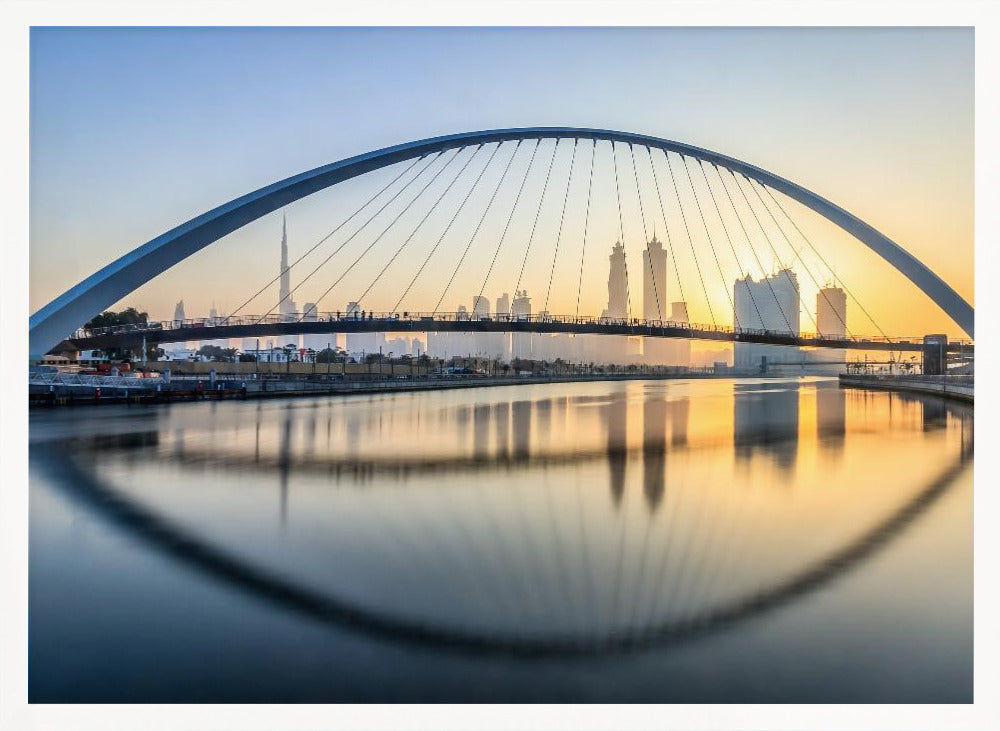A framed photograph of the Dubai Water Canal Bridge at sunrise, with its large arch spanning across the water. The Dubai skyline, including the Burj Khalifa, is visible in the background, silhouetted against a golden and blue sky. The bridge and buildings create a perfect reflection on the calm surface of the water. Wall Art