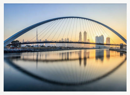 A framed photograph of the Dubai Water Canal Bridge at sunrise, with its large arch spanning across the water. The Dubai skyline, including the Burj Khalifa, is visible in the background, silhouetted against a golden and blue sky. The bridge and buildings create a perfect reflection on the calm surface of the water. Wall Art