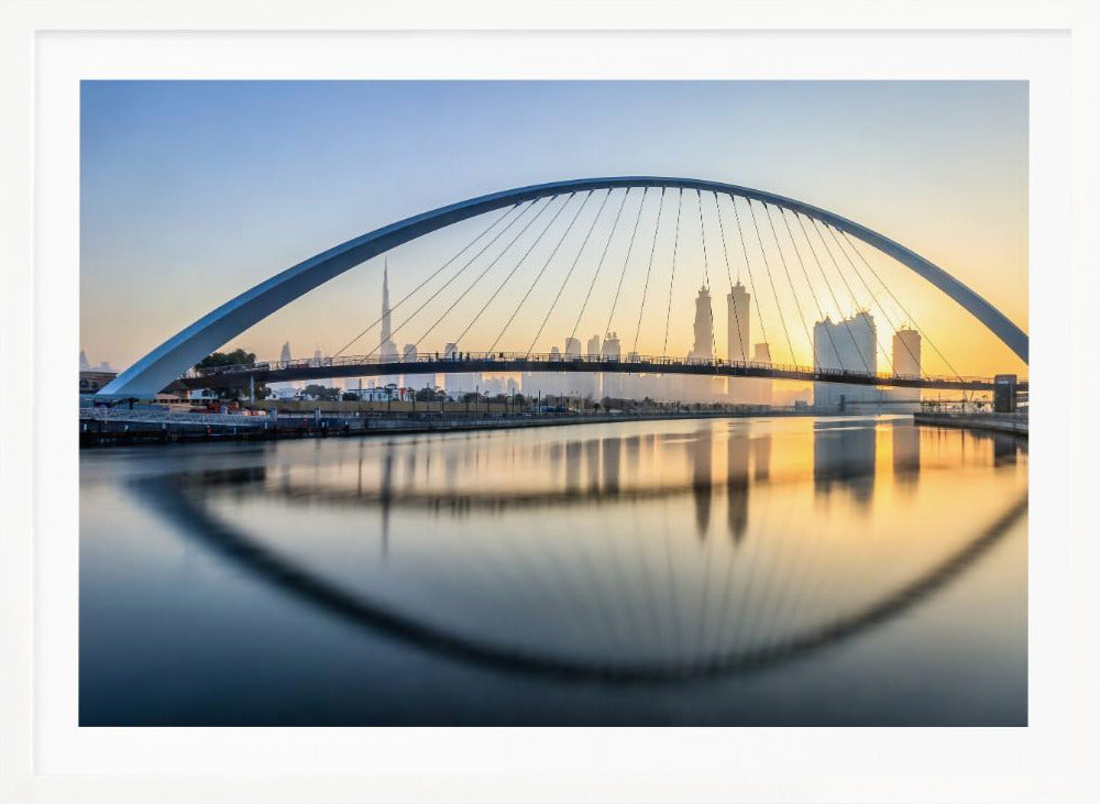 A framed photograph of the Dubai Water Canal Bridge at sunrise, with its large arch spanning across the water. The Dubai skyline, including the Burj Khalifa, is visible in the background, silhouetted against a golden and blue sky. The bridge and buildings create a perfect reflection on the calm surface of the water. Wall Art
