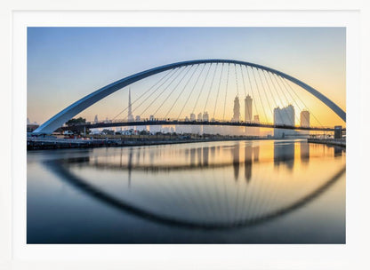 A framed photograph of the Dubai Water Canal Bridge at sunrise, with its large arch spanning across the water. The Dubai skyline, including the Burj Khalifa, is visible in the background, silhouetted against a golden and blue sky. The bridge and buildings create a perfect reflection on the calm surface of the water. Wall Art