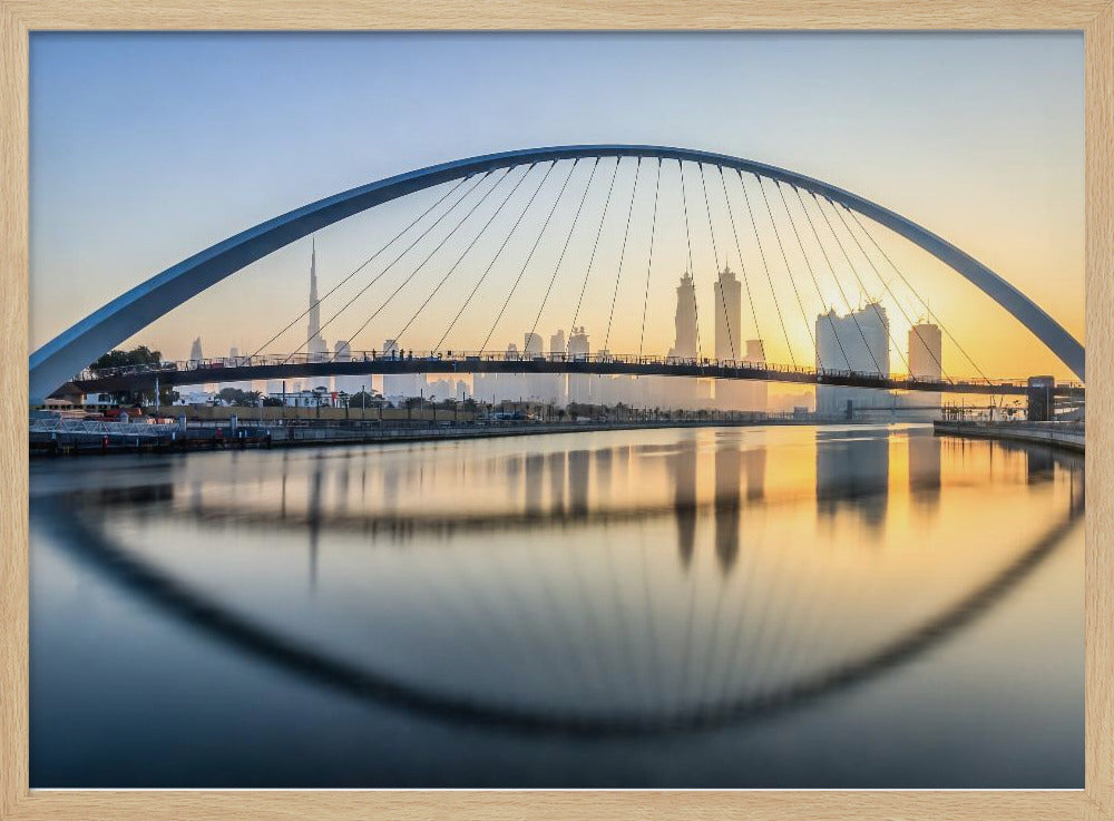 A framed photograph of the Dubai Water Canal Bridge at sunrise, with its large arch spanning across the water. The Dubai skyline, including the Burj Khalifa, is visible in the background, silhouetted against a golden and blue sky. The bridge and buildings create a perfect reflection on the calm surface of the water. Wall Art