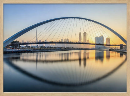 A framed photograph of the Dubai Water Canal Bridge at sunrise, with its large arch spanning across the water. The Dubai skyline, including the Burj Khalifa, is visible in the background, silhouetted against a golden and blue sky. The bridge and buildings create a perfect reflection on the calm surface of the water. Wall Art