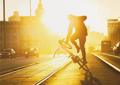 A silhouetted BMX rider performs a trick on a city street during a brilliant golden sunset, with the sun flaring behind them and casting long shadows on the tram tracks. Decor
