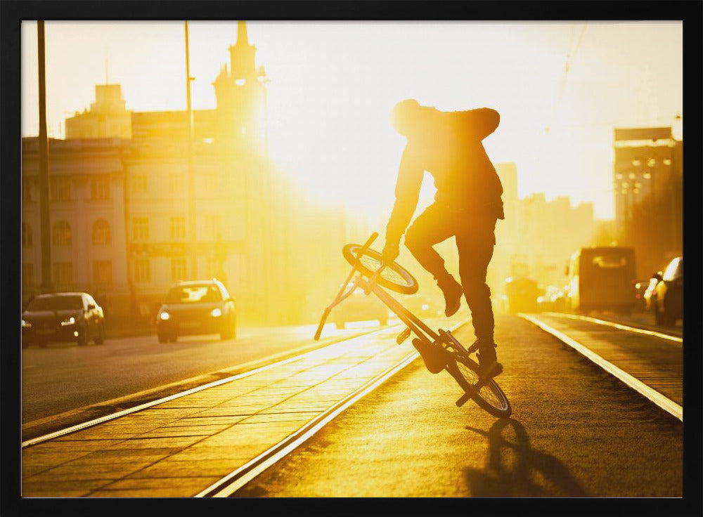 A silhouetted BMX rider performs a trick on a city street during a brilliant golden sunset, with the sun flaring behind them and casting long shadows on the tram tracks. Decor