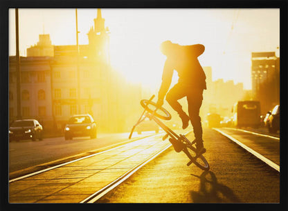A silhouetted BMX rider performs a trick on a city street during a brilliant golden sunset, with the sun flaring behind them and casting long shadows on the tram tracks. Decor