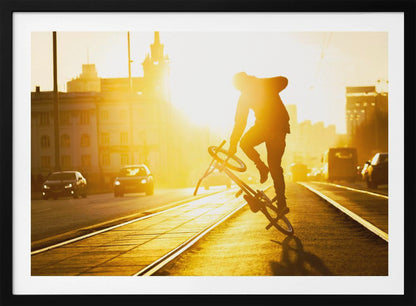 A silhouetted BMX rider performs a trick on a city street during a brilliant golden sunset, with the sun flaring behind them and casting long shadows on the tram tracks. Decor