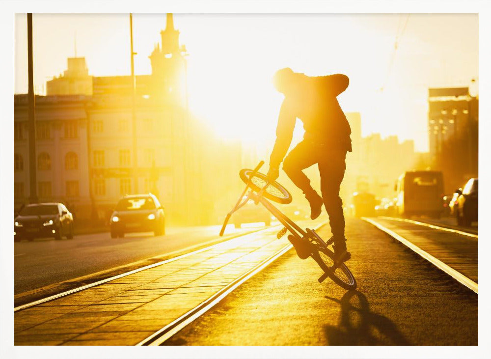 A silhouetted BMX rider performs a trick on a city street during a brilliant golden sunset, with the sun flaring behind them and casting long shadows on the tram tracks. Decor