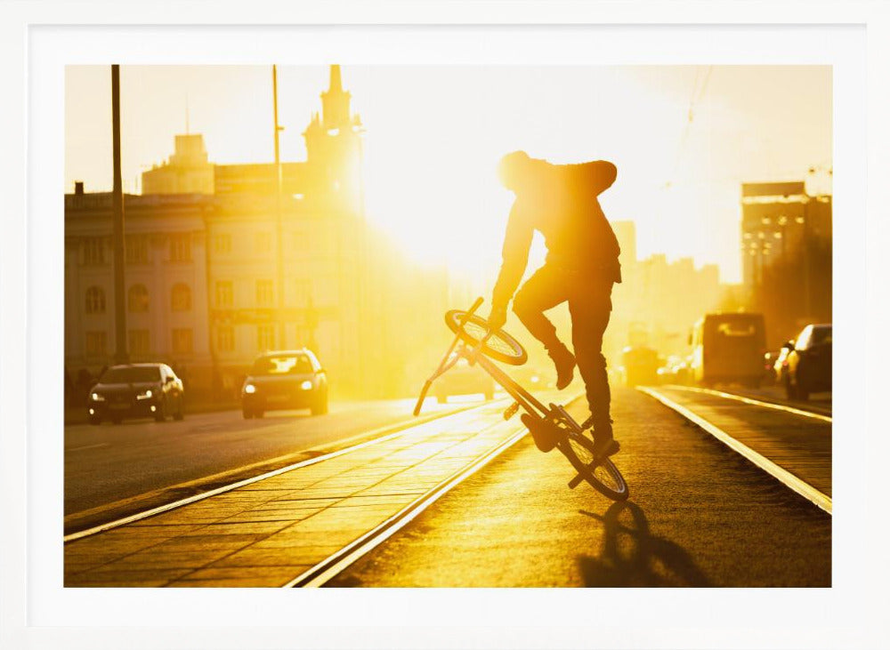 A silhouetted BMX rider performs a trick on a city street during a brilliant golden sunset, with the sun flaring behind them and casting long shadows on the tram tracks. Decor