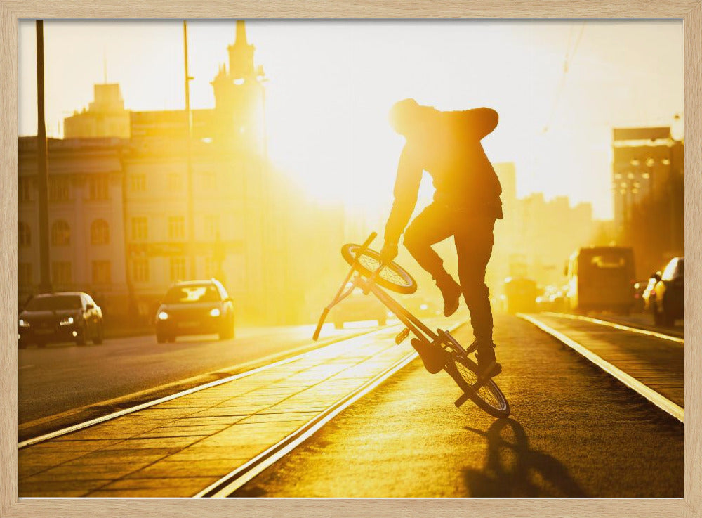 A silhouetted BMX rider performs a trick on a city street during a brilliant golden sunset, with the sun flaring behind them and casting long shadows on the tram tracks. Decor