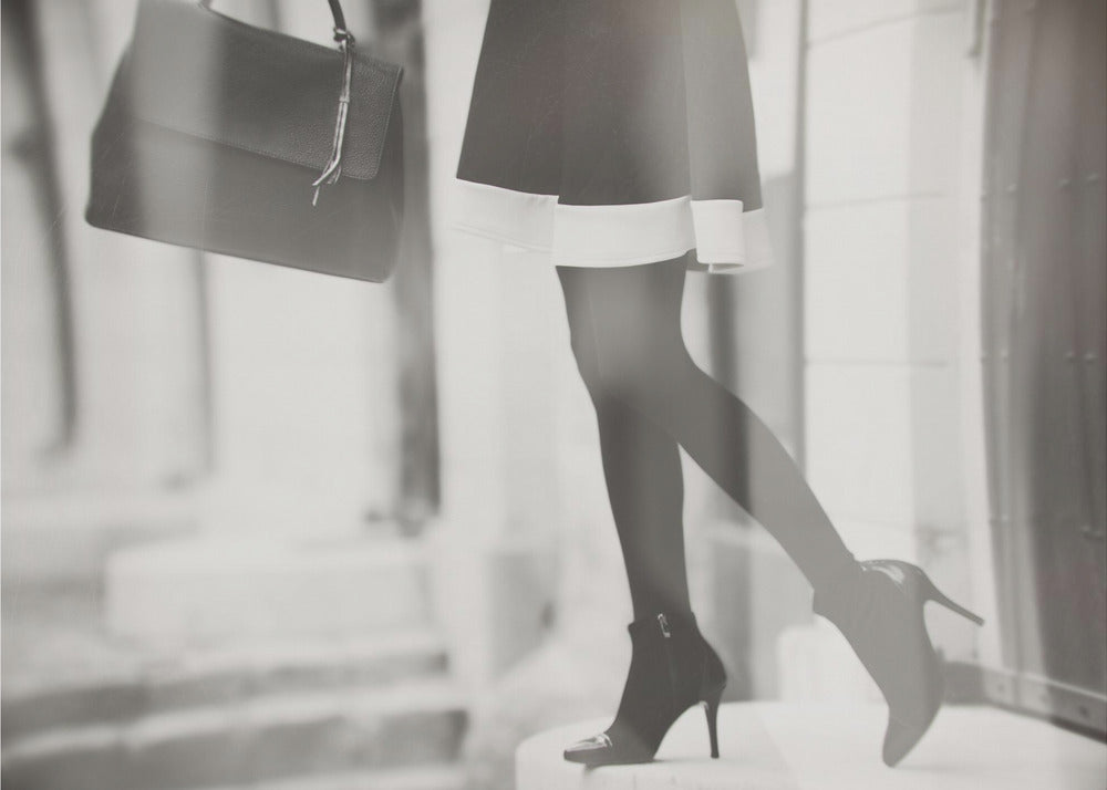 A framed, black and white fashion photograph focusing on the lower half of a woman as she walks. She is wearing a dark skirt with a white hem, black tights, and stylish high-heeled ankle boots, and she is carrying a dark handbag. Print
