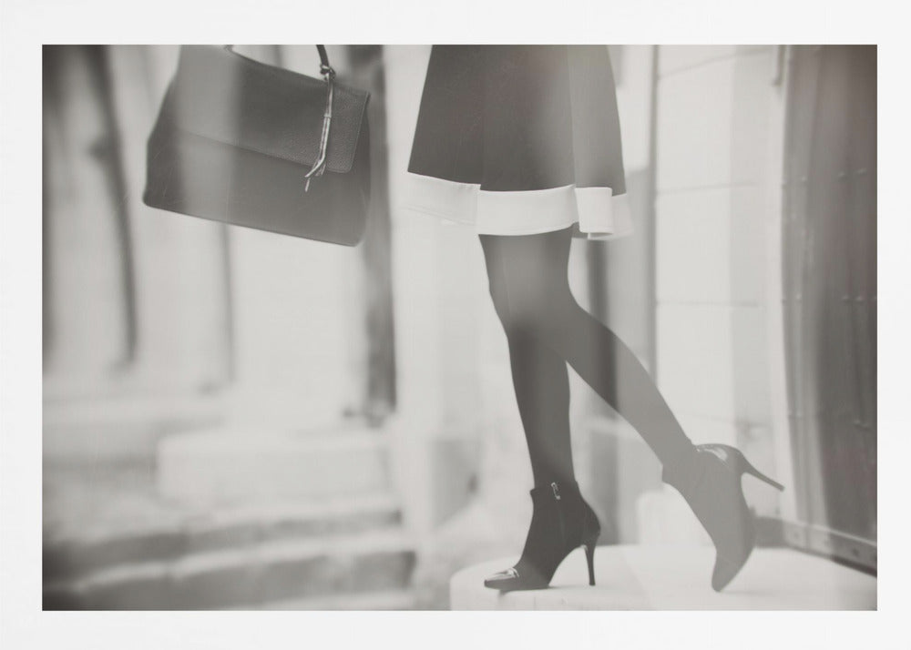 A framed, black and white fashion photograph focusing on the lower half of a woman as she walks. She is wearing a dark skirt with a white hem, black tights, and stylish high-heeled ankle boots, and she is carrying a dark handbag. Print