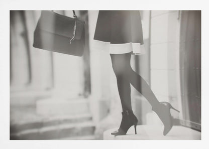 A framed, black and white fashion photograph focusing on the lower half of a woman as she walks. She is wearing a dark skirt with a white hem, black tights, and stylish high-heeled ankle boots, and she is carrying a dark handbag. Print