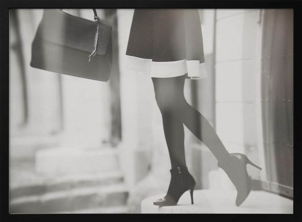 A framed, black and white fashion photograph focusing on the lower half of a woman as she walks. She is wearing a dark skirt with a white hem, black tights, and stylish high-heeled ankle boots, and she is carrying a dark handbag. Print