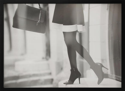 A framed, black and white fashion photograph focusing on the lower half of a woman as she walks. She is wearing a dark skirt with a white hem, black tights, and stylish high-heeled ankle boots, and she is carrying a dark handbag. Print