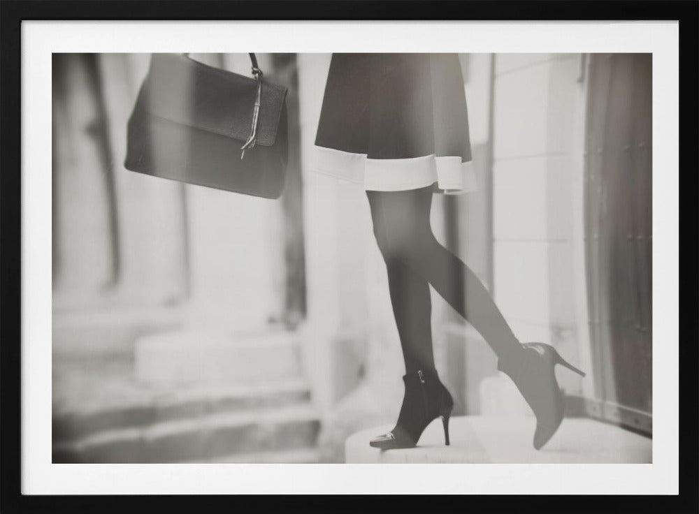 A framed, black and white fashion photograph focusing on the lower half of a woman as she walks. She is wearing a dark skirt with a white hem, black tights, and stylish high-heeled ankle boots, and she is carrying a dark handbag. Print