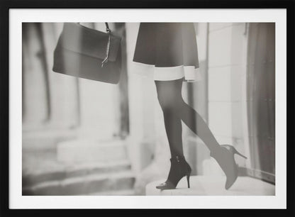 A framed, black and white fashion photograph focusing on the lower half of a woman as she walks. She is wearing a dark skirt with a white hem, black tights, and stylish high-heeled ankle boots, and she is carrying a dark handbag. Print
