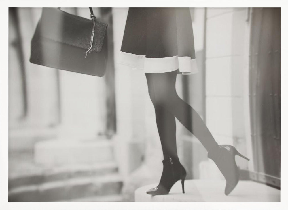 A framed, black and white fashion photograph focusing on the lower half of a woman as she walks. She is wearing a dark skirt with a white hem, black tights, and stylish high-heeled ankle boots, and she is carrying a dark handbag. Print