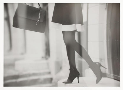 A framed, black and white fashion photograph focusing on the lower half of a woman as she walks. She is wearing a dark skirt with a white hem, black tights, and stylish high-heeled ankle boots, and she is carrying a dark handbag. Print