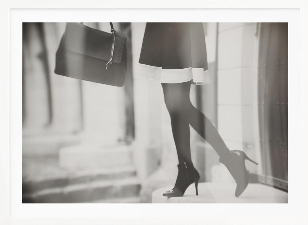 A framed, black and white fashion photograph focusing on the lower half of a woman as she walks. She is wearing a dark skirt with a white hem, black tights, and stylish high-heeled ankle boots, and she is carrying a dark handbag. Print